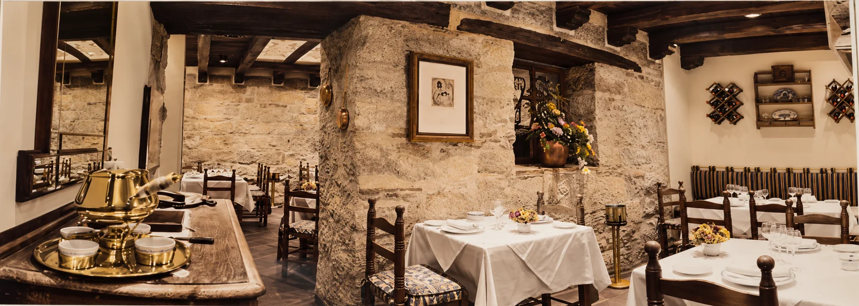 Panorámica del comedor de La Aldaba: muro de piedra, vigas de madera, cuadros y mesas con mantel blanco